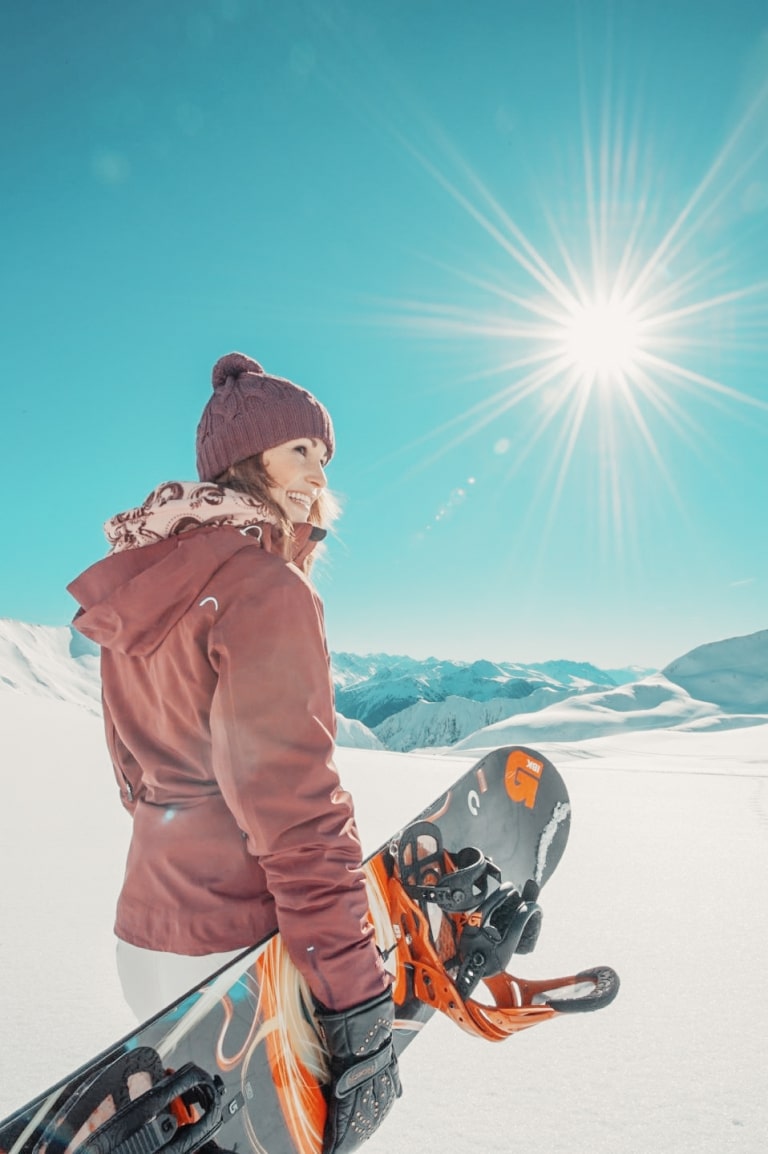 Frau hält Snowboard und schaut in Richtung Kamera
